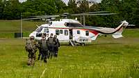 Soldaten in Uniform und zivil gekleidete Personen laufen hintereinander über eine Wiese zu einem Hubschrauber