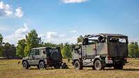 Zwei militärische Fahrzeuge stehen in einer Heidelandschaft. Um sie herum stehen beziehungsweise liegen bewaffnete Soldaten.