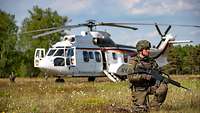 Ein weißer Hubschrauber steht auf einer Wiese. Vor ihm knieen bewaffnete Soldaten und sichern ihn.