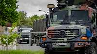Militär-LKW bewegen sich in Kolonne durch eine ländliche, frühlingshafte Landschaft.