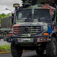 Militär-LKW bewegen sich in Kolonne durch eine ländliche, frühlingshafte Landschaft.