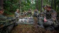 Mehrere Soldaten im Wald knien im Kreis um eine Plane, auf der Kartons und Tüten liegen.