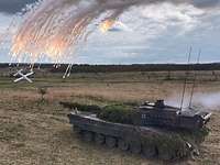 Eine Drohne fliegt auf einen fahrenden Panzer zu. Leuchtmunition ist in der Luft zu sehen.
