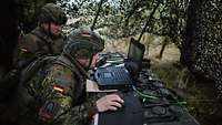 Zwei Soldaten sitzen vor einem Laptop unter einem Tarnnetz im Gelände.
