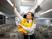 Eine Lufttransportbegleiterin steht im Flugzeug und führt vor, wie man eine Rettungsweste anlegt.