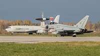 Ein großes AWACS-Aufklärungsflugzeug fährt hinter einem Eurofighter-Kampfjet auf der Air Base Ämari.
