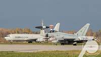 Ein großes AWACS-Aufklärungsflugzeug fährt hinter einem Eurofighter-Kampfjet auf der Air Base Ämari.