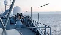 Ein Soldat steht mit einer Waffe auf der Schulter an Deck eines Schiffes und feuert ein Geschoss ab.
