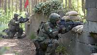 Zwei Soldaten knien mit Waffe hinter grauen Deckungen im Wald.