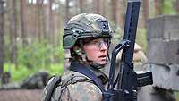 Eine Soldatin kniet mit dem Gewehr hinter einer grauen Deckung im Wald.
