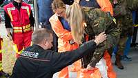 Ein Feuerwehrmann hilft einer Soldatin beim anlegen des Schutzanzug, eine weitere Frau unterstützt.