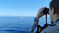 Eine Soldatin im Irini-Einsatz beobachtet mit einem Fernglas einen Frachter im Mittelmeer