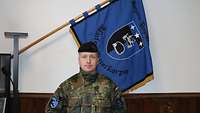 Ein Soldat mit einem schwarzen Barett steht vor einer blauen Fahne mit einem Wappen darauf.