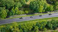 Eine Straße aus der Vogelperspektive, auf der 4 Autos fahren. Neben der Straße Wiesen und Wald.