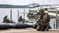 2 bewaffnete Soldaten mit Funkgeräten knien auf dem Boden, im Wasser 2 Boote mit Soldaten.