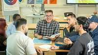 Ein Mann mit Buch in der Hand sitzt mit jungen Erwachsenen an einem Tisch und lächelt in die Kamera.