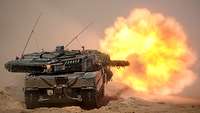 Ein Panzer schießt auf einer sandigen Freifläche. Ein heller Feuerball wird sichtbar.