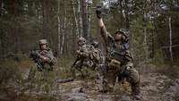 Mehrere Soldaten knien im Wald. Ein Soldat hält eine Drohne hoch.