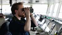 Ein Soldat steht auf der Kommandobrücke eines Schiffes und schaut durch ein Fernglas.