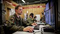 Drei Soldaten sitzen vor Computerbildschirmen an Tischen in einem hellen holzverkleideten Raum.