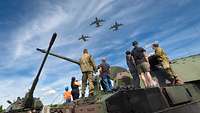 Personen stehen auf einer Panzerhaubitze 2000 und beobachten den Überflug von drei Transportflugzeugen