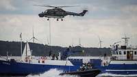 Über einem Schiff fliegt ein Hubschrauber und vor dem Schiff ist ein Schnellboot zu sehen.
