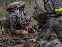 Zwei Soldaten knien vor einem liegenden Verwundeten auf Waldboden und leisten Erste Hilfe.