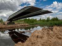Ein Panzer legt eine Verlegebrücke über einen Fluss am sandigen Ufer.