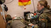 Eine Soldatin versorgt einen auf einer Liege liegenden Soldaten mit Bandagen um den Arm. 