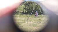 Durch eine Zieloptik mit Fadenkreuz ist ein Zahlenschild zu erkennen, das an einem Gestell hängt.