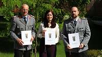 Zwei Soldaten und eine Frau stehen nebeneinander. Alle halten ein Dokument in die Kamera.