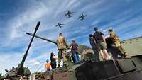 Personen stehen auf einer Panzerhaubitze und beobachten den Überflug von drei Transportflugzeugen.