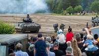 Mehrere Zuschauer schauen sich eine Vorführung mit Panzern und Soldaten im Gelände an.