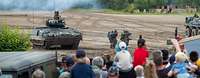Mehrere Zuschauer schauen sich eine Vorführung mit Panzern und Soldaten im Gelände an.