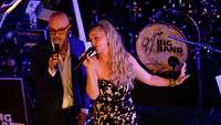 Ein Mann mit Brille im Anzug und eine blonde Frau im Kleid singen gemeinsam auf einer Bühne.