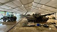 In einem großen Zelt stehen im Vordergrund ein Panzer und ein Teleskoplader. 