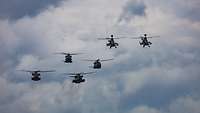 Sechs Hubschrauber fliegen in Formation vor blau-weißem Himmel. 