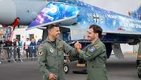 Zwei Piloten in Fliegerkombi geben sich lachend die Hand. Im Hintergrund ein Eurofighter mit Jubiläumsfolierung. 