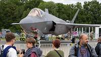 Ein graues Kampfflugzeug des Typs F-35A steht hinter einem Zaun. Davor stehen Besucher mit Kameras. 