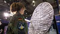 Soldatin in Flecktarn mit einem Patch des Weltraumkommando steht vor einem großen, silbernen Satellit. 