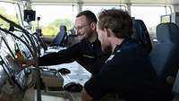 Two soldiers are sitting on the bridge of a ship, operating equipment.
