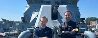 A German and a British soldier stand side by side on the deck of a military ship and smile.