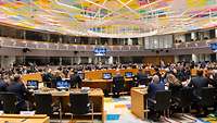 Ein großer Plenarsaal mit einem runden Tisch in der Mitte an dem Menschen aus Militär und Politik sitzen