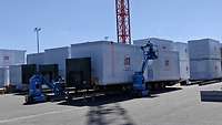 Mehrere verpackte Holzmodule stehen auf einem Platz und werden auf Anhänger verladen.