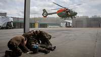 Ein Hubschrauber landet auf einem Platz. Im Vordergrund vier Soldaten. Sie schützen sich vor dem Wind.