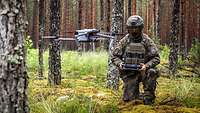 Ein Soldat kniet im Wald und steuert eine Drohne.