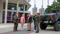 Fünf Personen, militärisch und zivil, stehen vor einem Gebäude. Rechts im Bild steht ein militärisches Fahrzeug.