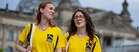 Zwei junge Frauen in gelben TShirts gehen nebeneinander. Im Hintergrund ist der Bundestag zu sshen.
