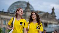 Zwei junge Frauen in gelben TShirts gehen nebeneinander. Im Hintergrund ist der Bundestag zu sshen.