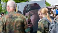 eine Bilderausstellung auf einem Fest zeigt ein übergroßes Porträt eines im Gesicht vernarbten Soldaten